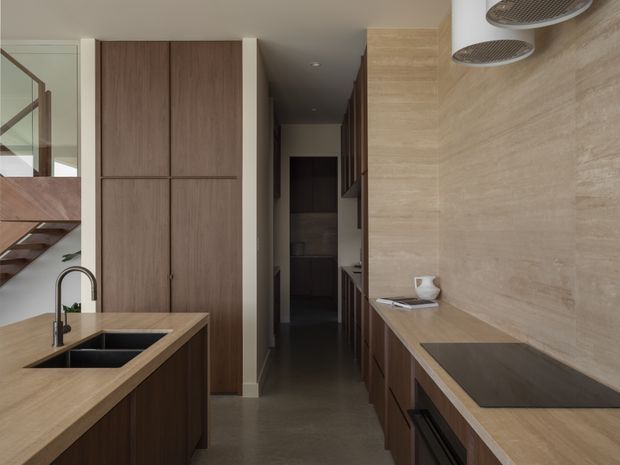 A narrow contemporary kitchen with dark wood cabinetry, light timber-toned benchtops, and a matching textured stone or tile backsplash. The space includes a central island with an undermount double sink and black tapware, integrated cabinetry and appliances along the right wall, and a hallway or pantry zone beyond. A glazed partition and staircase are visible at left.