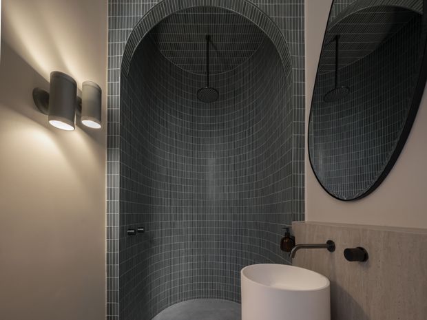 A moody contemporary bathroom featuring a curved walk-in shower lined with dark grey small-format tiles, a round overhead shower, warm wall lighting, a freestanding white basin, and a stone-look vanity backsplash with black tapware. A large oval mirror reflects the shower enclosure.
