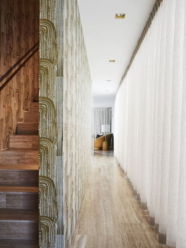 A long, bright interior hallway with a timber staircase on the left, dramatic patterned wall finishes in brown, olive and cream, a textured pale wall on the right, recessed ceiling lights, and light timber flooring. A small seating or living area is visible at the far end.