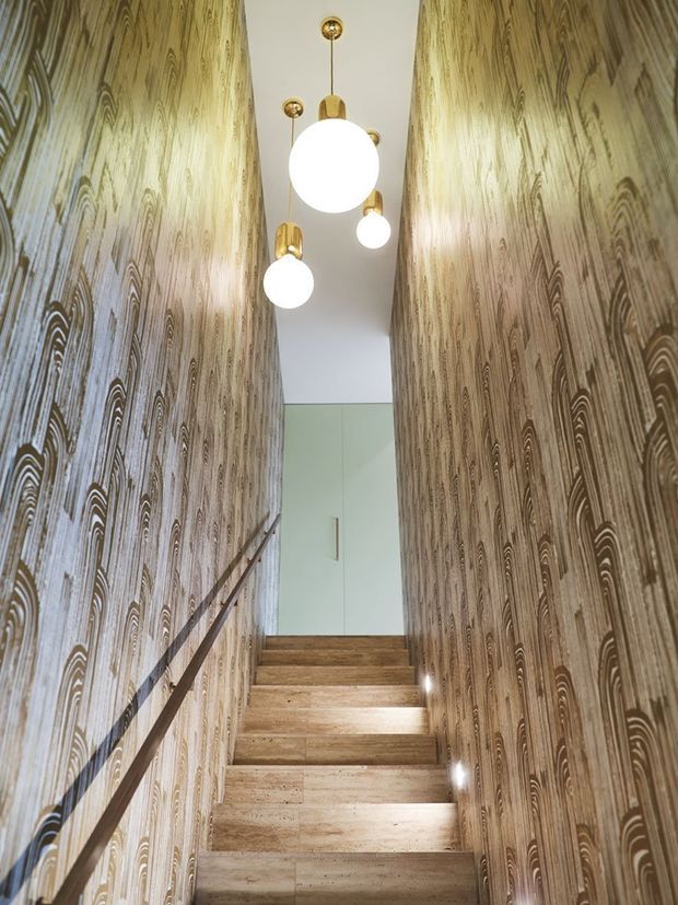 A narrow interior stairway rises between two walls covered in dramatic brown and beige patterned wallpaper with a wood-grain-like motif. The stairs have a warm timber finish, a slim dark handrail runs along the left wall, and several spherical pendant lights with brass-toned fittings hang from the white ceiling. A pale built-in door or cupboard is visible at the landing.