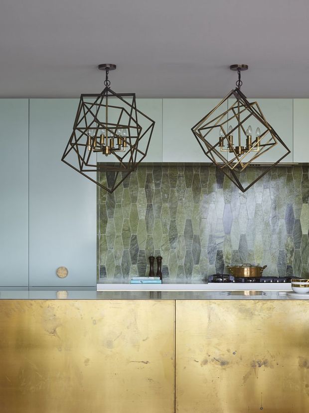 Close-up view of a sophisticated kitchen with a large polished gold-toned metal island or cabinetry front, pale blue-grey cabinetry, and a striking irregular green-grey stone backsplash. Two oversized geometric dark metal pendant chandeliers hang above the counter, creating a dramatic, luxurious focal point.