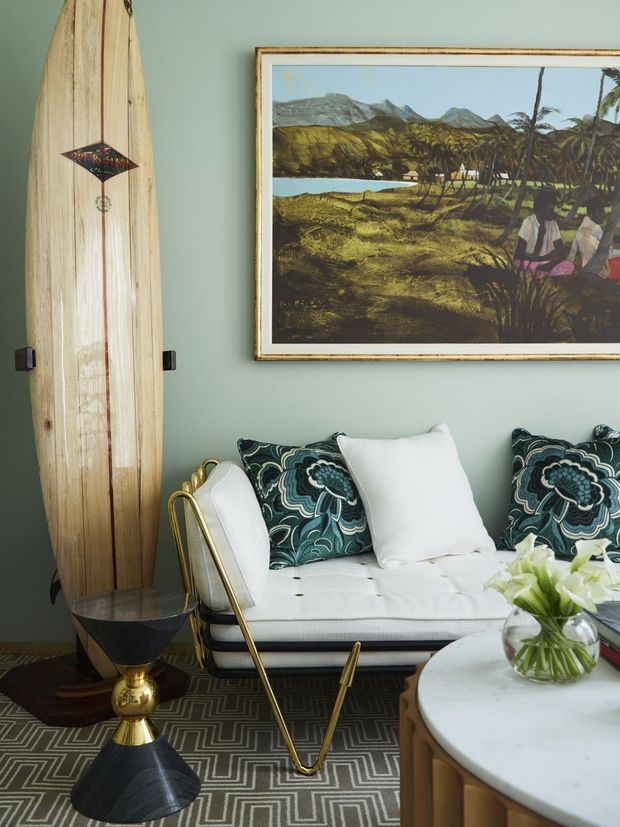 A richly styled living room vignette with a white upholstered sofa, patterned teal cushions, brass detailing, a marble-topped coffee table, and a vintage surfboard displayed vertically against a muted sage-green wall. A framed tropical landscape artwork and geometric rug add character and pattern.