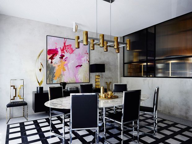 A sophisticated contemporary dining room with a large round marble dining table surrounded by black upholstered chairs. The space features a dramatic geometric black-and-white floor, textured grey concrete-look walls, a ribbed white ceiling, black-framed glazed cabinetry or partitioning, and a sculptural brass pendant chandelier. Abstract artwork and brass accents add a luxurious, high-contrast character.