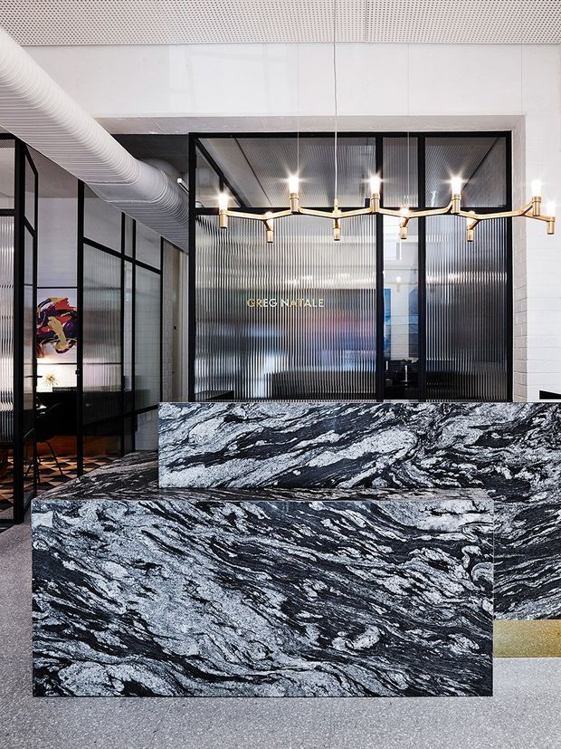 A polished commercial reception area with a dramatic black-and-white veined stone reception desk and stepped counter. Black-framed glazed partitions with ribbed translucent glass enclose offices behind it, while exposed white ductwork, a perforated ceiling, and a linear brass pendant chandelier create an industrial contemporary fit-out. Terrazzo-like flooring and white masonry walls complete the space.