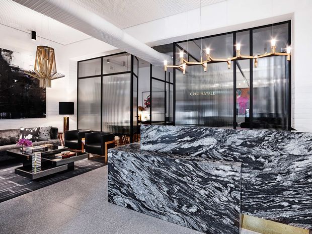 A sophisticated contemporary commercial reception or hospitality interior with a dramatic black-and-white marble reception counter in the foreground. Behind it are black-framed fluted-glass partitions, a lounge seating area, sculptural pendant lighting, dark artwork, and polished black and brass accents. The space has a bright, refined, high-contrast aesthetic with industrial-style glazing and premium stone finishes.