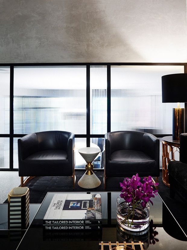 A sophisticated modern sitting area with two black leather lounge chairs positioned in front of tall fluted-glass windows framed in black metal. A sculptural white and brass side table sits between the chairs, while a black glass coffee table holds design books and a clear vase of purple orchids. The room has a textured grey plaster ceiling or upper wall, dark flooring, dramatic black furnishings, and warm metallic accents.