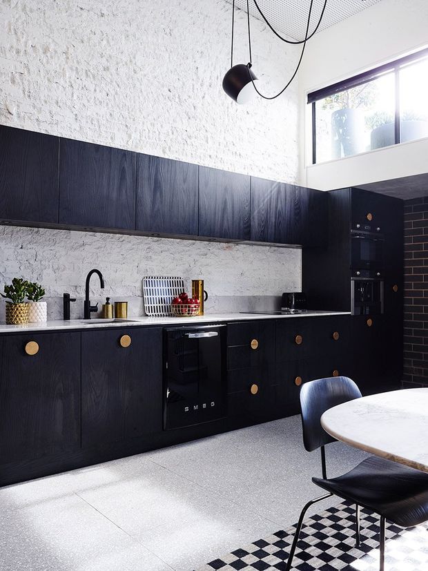 A dramatic contemporary kitchen with full-height black timber cabinetry, round brass knobs, integrated appliances, and a pale stone benchtop. White-painted textured brick walls contrast with the dark joinery, while a sculptural black pendant hangs from a perforated ceiling. A small dining table and black upholstered chairs sit in the foreground beside a monochrome checkerboard rug, with clerestory glazing bringing in natural light.