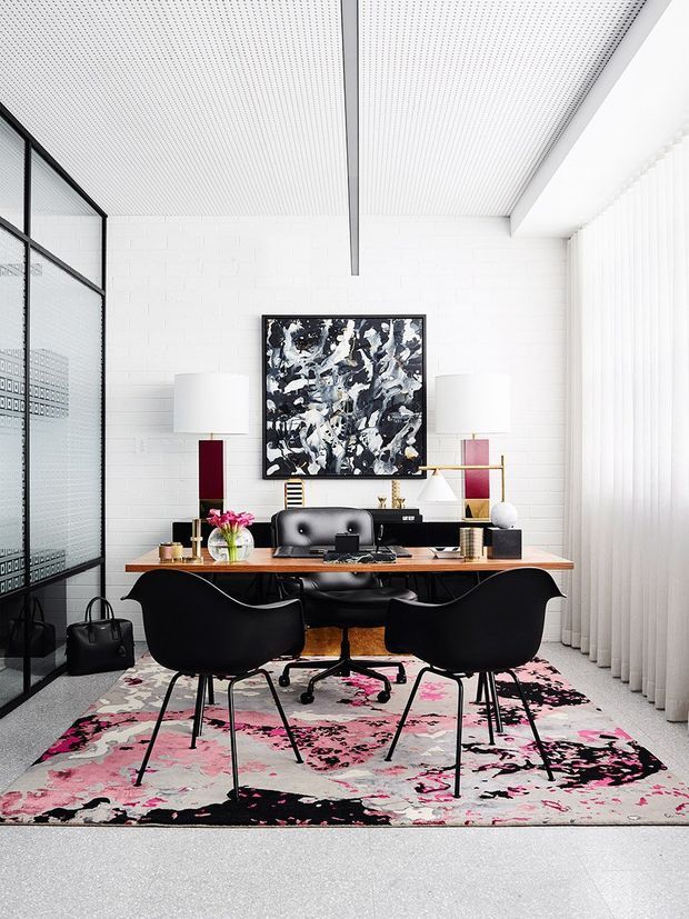 Bright, high-contrast home office with a long timber desk, dual workstations, black task chairs, sculptural black guest chairs, and a bold pink, black, and white patterned rug. The room has white painted brick walls, a white perforated ceiling with a dark exposed beam, full-height sheer curtains, black-framed glazed partitioning, contemporary artwork, and restrained decorative objects.