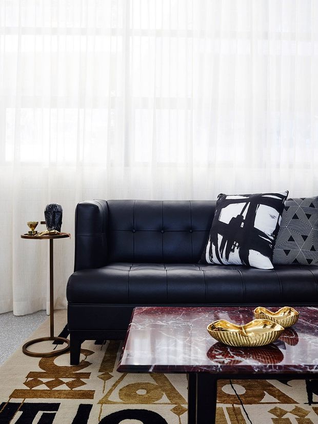 A stylish living room vignette centered on a black tufted leather sofa, with bold black-and-white patterned cushions, sheer white curtains, a geometric area rug, and a glossy burgundy marble coffee table topped with sculptural gold bowls. The space has a refined, high-contrast contemporary look with natural daylight.