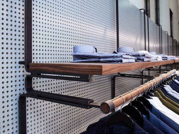 Close-up of a contemporary retail clothing display system, with folded garments on a dark timber shelf and hanging clothing below. The display is mounted against a perforated light-grey metal wall panel with black metal brackets and rails.