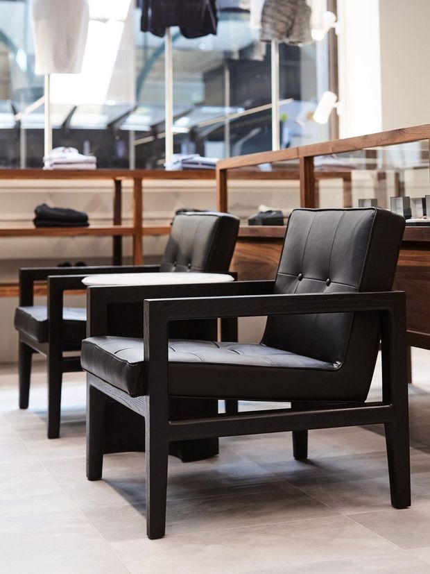 A refined retail showroom interior displaying black leather lounge chairs with dark timber frames. Large mirrors and glass display cabinetry line the background, with timber shelving and folded clothing visible. The space has a bright, polished, contemporary feel and pale tiled flooring.