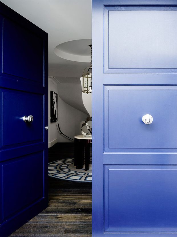 A dramatic view through open deep-blue paneled doors into a refined residential entry or hallway. The space beyond features dark timber flooring, a geometric circular rug, a curved white ceiling detail, a console table with decorative objects, and a hanging glass light fixture. The image has a bold blue-and-white palette with polished traditional detailing.