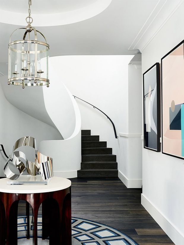 Elegant residential entry hall with a dramatic curved staircase, white sculptural balustrade, black stair treads, dark timber flooring, and a slim black handrail. A chrome sculptural object sits on a round console table beneath a glass-and-metal pendant chandelier. Framed abstract artwork lines the white wall, while a patterned blue, white, and black rug adds contrast. The space has a refined, contemporary interior with strong monochrome geometry and Art Deco-inspired detailing.