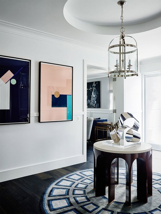 A refined, light-filled interior foyer or hallway with white paneled walls, a dark timber floor, geometric patterned rug, framed abstract artwork, and a sculptural mirrored object displayed on a round dark wood console. A large circular recessed ceiling detail and glass-and-metal pendant light add architectural interest. An adjoining room is visible through a wide opening.