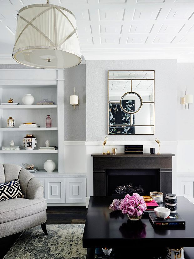 Elegant, light-filled living room with a black fireplace surround, built-in white shelving and cabinetry, a large decorative mirror, tailored grey seating, and a glossy black coffee table. The room features a coffered white ceiling, geometric chandelier, monochrome palette, and refined gold and black accents.