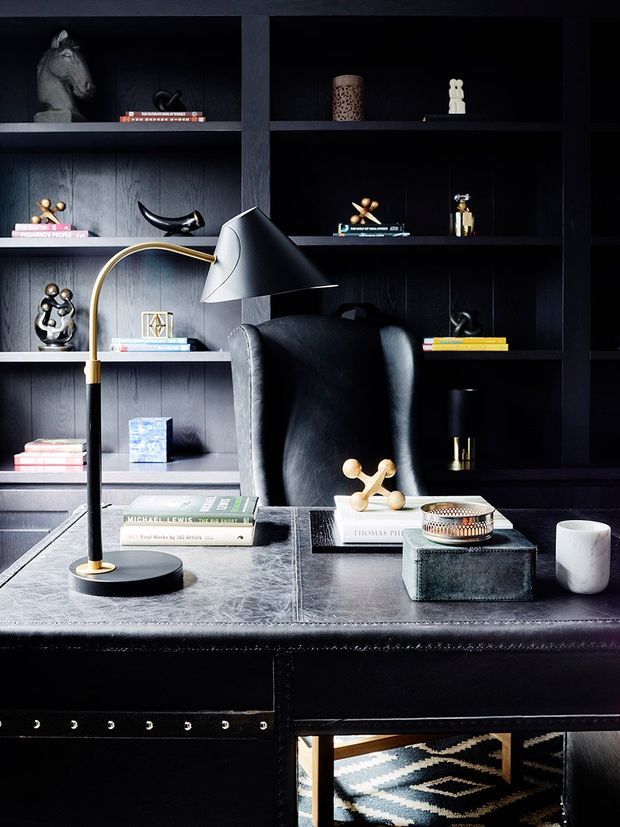 A moody, refined home office with dark built-in shelving, a black leather high-back chair, black stone or leather-topped desk, and a sculptural brass-and-black desk lamp. The shelving displays books and small decorative objects, while a patterned rug is partially visible below.