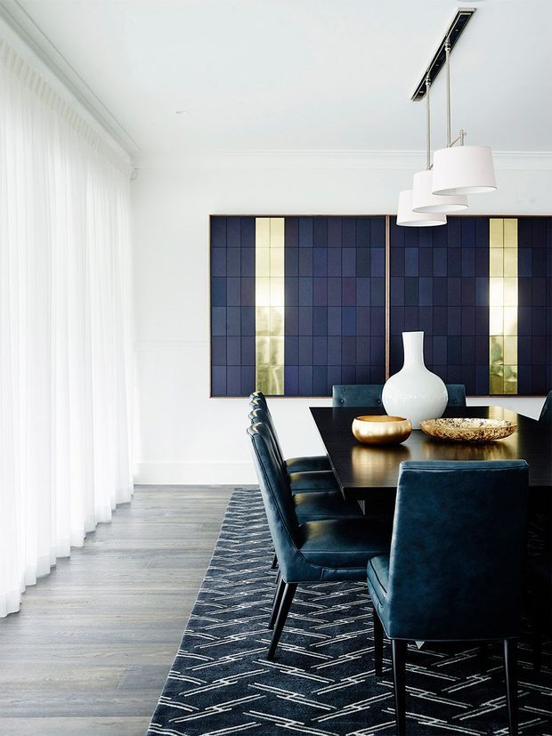 Bright, refined dining room with a long dark dining table surrounded by deep navy upholstered chairs. A large geometric navy-and-gold tiled feature wall provides a bold backdrop, complemented by sculptural white pendant lights, sheer floor-to-ceiling curtains, pale timber flooring, and a patterned dark rug. The space combines crisp white architectural detailing with rich, luxurious accents.