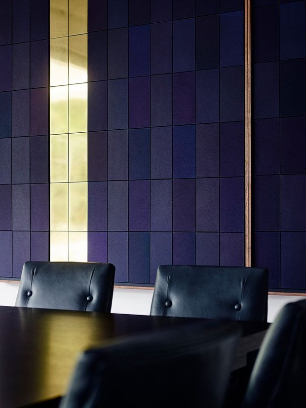 A dark, refined dining or meeting room focused on a large dark wood table and black leather button-tufted chairs. The backdrop is a dramatic grid of deep navy and purple rectangular wall panels, intersected by a narrow illuminated vertical feature and framed with warm timber trim. The scene has low, moody lighting and strong contrast, with a polished contemporary feel.