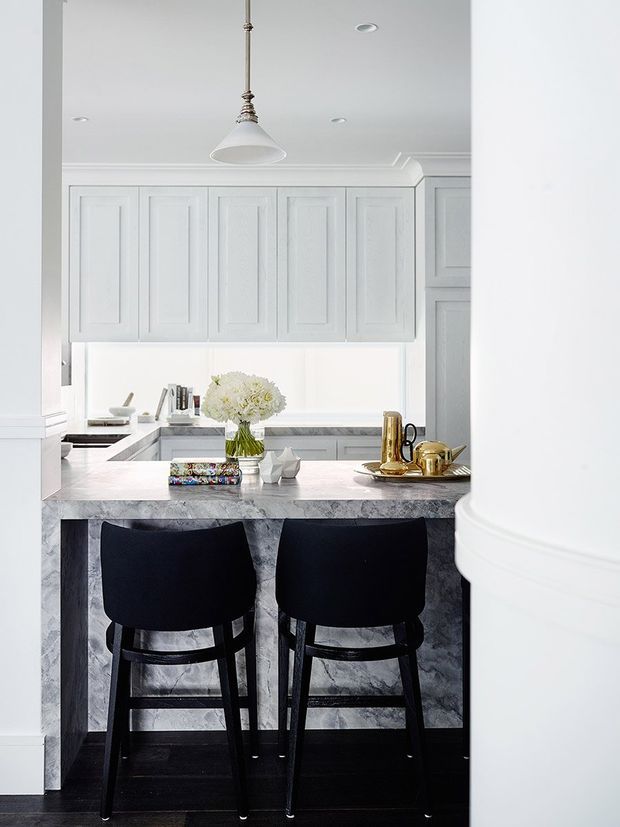Bright, refined kitchen viewed through a wide opening, featuring white shaker-style cabinetry, a grey veined stone island with a raised breakfast bar, two black upholstered bar stools, a pale stone benchtop, and a white pendant light. Decorative books, flowers, ceramics, and gold-toned serving pieces add restrained accents. White architectural trim and the black floor create a crisp, high-contrast composition.