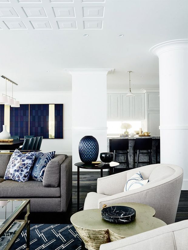 Bright, refined open-plan living room with a formal white paneled ceiling, substantial decorative columns and mouldings, grey upholstered seating, patterned blue-and-white cushions, a dark geometric rug, sculptural side tables, and a blue feature artwork. A contemporary kitchen with dark cabinetry and a stone island is visible in the background.