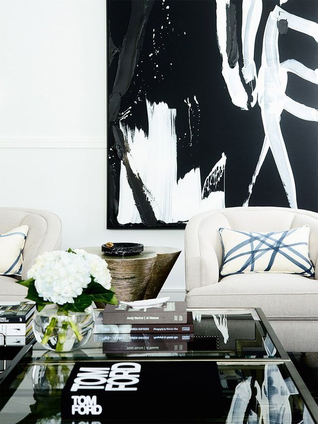 A refined living room vignette with two cream upholstered armchairs facing a glossy black glass coffee table. The table holds stacked design books, a clear vase of white hydrangeas, and a metallic decorative bowl. A large black-and-white abstract artwork forms a dramatic feature behind the seating, with blue-and-white patterned cushions adding contrast. The space is bright, polished, and contemporary with strong monochrome styling.