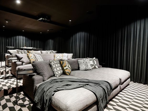 A dark, sophisticated media room with a large grey sectional sofa and chaise, layered patterned cushions, and a charcoal throw. Full-height black curtains line the walls, while a bold black-and-white geometric carpet creates strong visual contrast. Recessed ceiling lighting and a wall-mounted screen area reinforce the cinematic atmosphere.