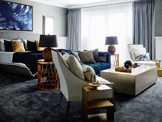 Elegant, richly layered living room with a deep blue sofa, white and charcoal upholstered seating, patterned cushions, sculptural table lamps, a large square metallic coffee table, and dark plush carpeting. Floor-to-ceiling sheer and grey drapery fills the bright window wall, while grey textured wallcovering, framed abstract artwork, and warm metallic occasional tables create a refined, contemporary atmosphere.