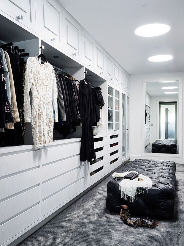 Bright, highly organised walk-in wardrobe with extensive white built-in cabinetry, open hanging rails, drawers, glass-front storage and integrated lighting. A large black tufted ottoman sits centrally on a grey carpet, with a full-height mirror at the end of the room reflecting additional wardrobes and a doorway.