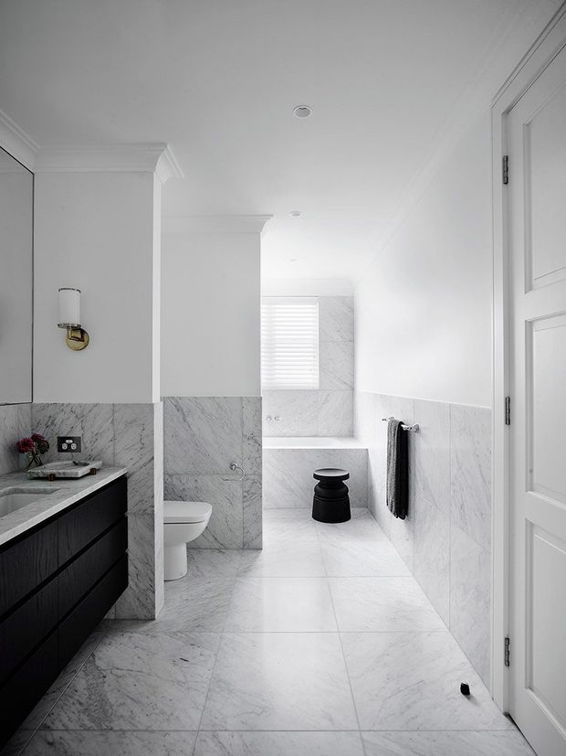 A bright, refined bathroom viewed from the doorway, featuring full-height white marble-look wall and floor tiling, a dark timber vanity with pale stone benchtop, white toilet, freestanding bathtub at the rear, black stool, and black-and-white fixtures. Classic wall panelling, moulded cornices, and a paneled door add traditional detailing.