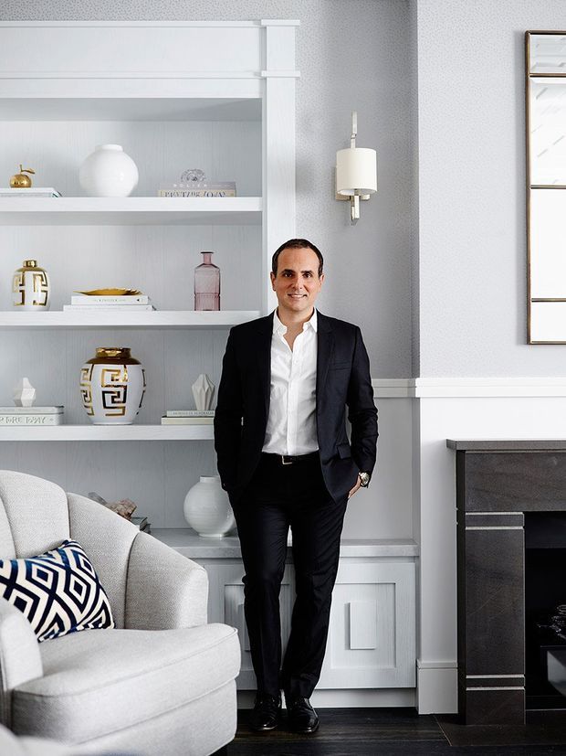A refined living room interior with pale grey built-in shelving displaying decorative objects and books, a light upholstered armchair with a bold geometric cushion, a dark fireplace surround, white wall panelling and trim, and a contemporary wall sconce. A formally dressed man stands in the centre of the room.