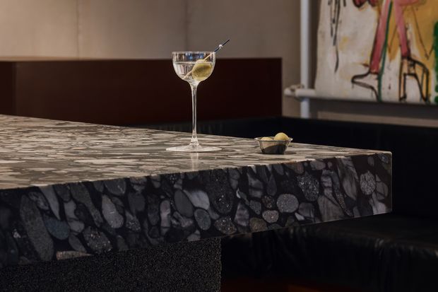 Close-up of a sophisticated bar or dining counter with a dark, dramatically patterned stone surface and thick waterfall-style edge. A cocktail and small snack sit on the counter, with dark upholstered seating and colourful abstract artwork in the background. The atmosphere is intimate, moody and refined.