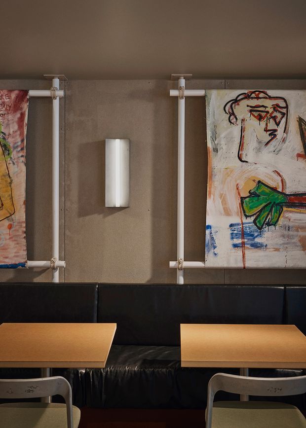 A moody commercial dining interior with black upholstered banquette seating, light timber tables, and pale metal chairs. The taupe wall features exposed white conduit pipes, a simple wall sconce, and colourful abstract artwork displayed behind the seating.