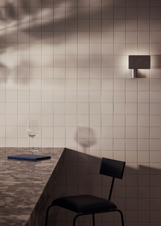 Moody close-up of a minimalist dining or kitchen corner featuring a pale square-tiled wall, a dark wall-mounted rectangular light, a marble-look stone countertop, a wine glass, books, and a black modern chair. Strong directional lighting creates dramatic shadows across the tiled surface.
