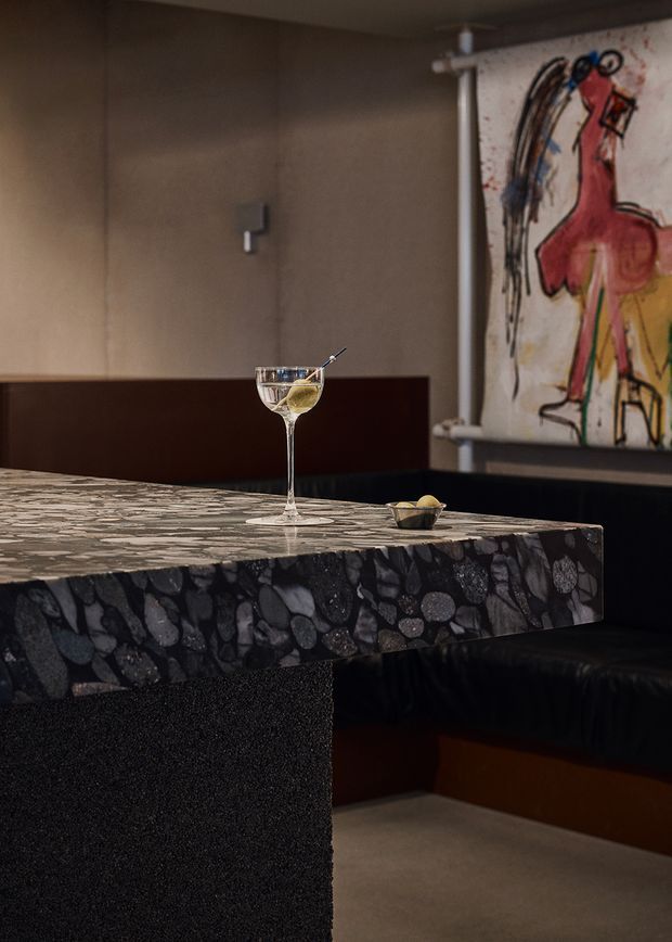 A moody, refined bar or dining nook featuring a thick dark terrazzo countertop with exposed rounded stone aggregate, supported by a textured dark pedestal. A glass of wine and small bowl sit on the counter, with a black upholstered banquette behind it. The space has warm taupe walls, dark timber or painted wall panels, and a large expressive abstract artwork mounted on a scroll-like frame.