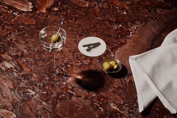 Close-up styled tabletop scene featuring a richly veined dark reddish-brown stone surface, a cocktail glass with an olive, a small bowl of olives, a round coaster, and a white folded napkin. The image highlights the dramatic natural pattern and polished finish of the table.