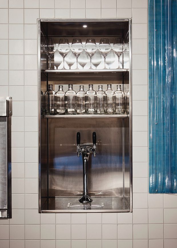 A compact built-in stainless steel bar niche set into a white square-tiled wall. The recessed shelving displays wine glasses and clear bottles, with a polished beer tap mounted on the lower shelf. A vivid blue ribbed glass or acrylic panel borders the niche on the right, adding a strong accent. The scene has a clean, utilitarian hospitality feel with reflective metal surfaces.