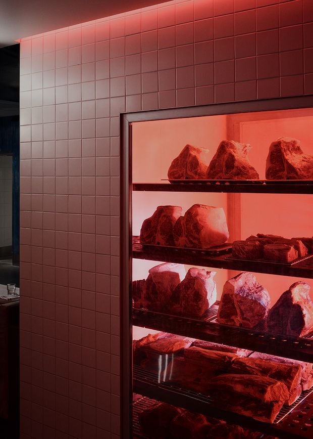A close-up interior view of a commercial meat display or butchery space. Large cuts of meat are arranged on multiple shelves inside a red-lit refrigerated display cabinet, set against a wall of square pinkish-red tiles. The scene has a dramatic, industrial retail atmosphere.