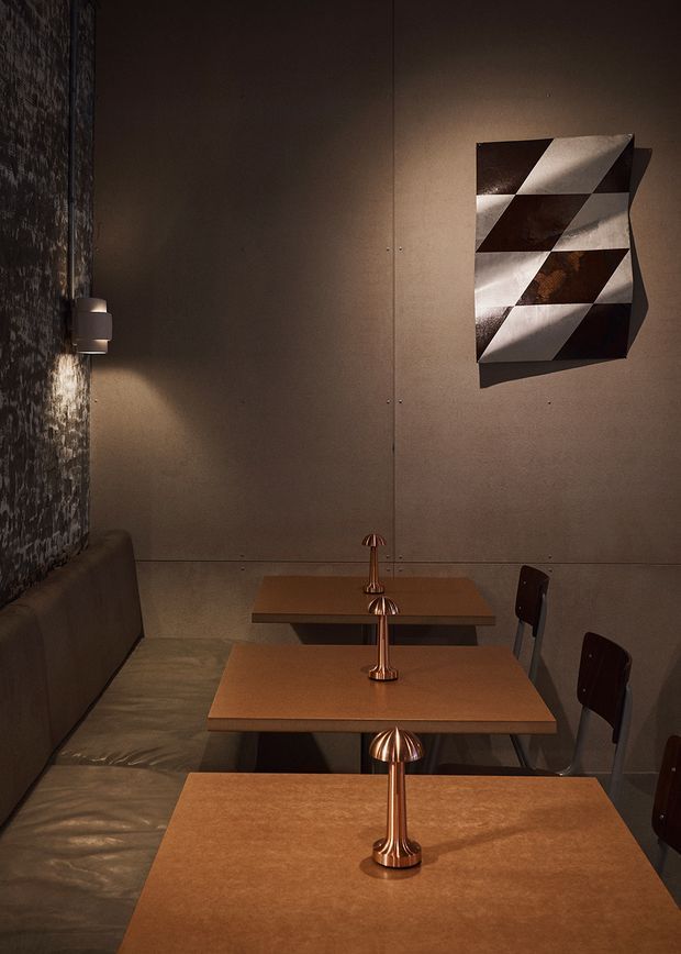 A dimly lit intimate dining area with a long upholstered banquette along one wall and several small square timber tables arranged in a row. Copper-toned table lamps sit centrally on each table, while dark dining chairs line the opposite side. The room has dark textured wall finishes, warm brown tones, and a geometric monochrome wall artwork, creating a moody, refined restaurant-like atmosphere.