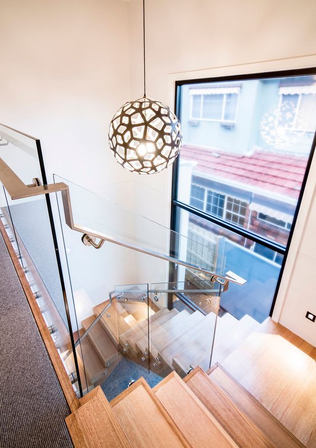 A bright, contemporary stairwell viewed from above, featuring a timber staircase with clear glass balustrades, slim metal handrails, and a large black-framed window. A sculptural spherical pendant light hangs over the stair void, with neighbouring buildings visible outside.