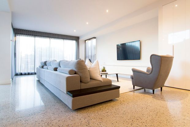 Bright contemporary living room with a large pale sectional sofa, sculptural armchair, low dark timber coffee table, wall-mounted television, floor-to-ceiling sheer curtains, glossy white built-in cabinetry, recessed ceiling lights, and a polished light terrazzo floor. The space has abundant natural light and a clean, open layout.