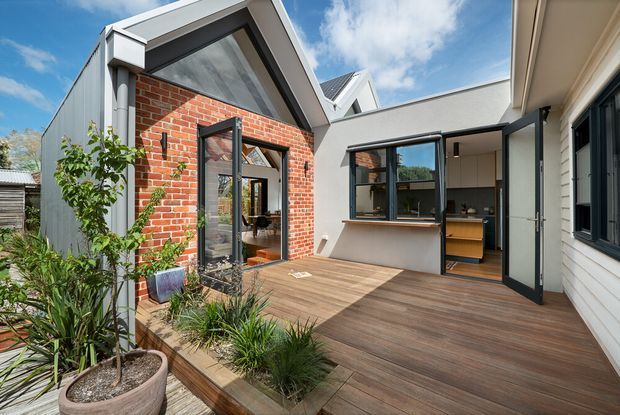 A bright contemporary courtyard-style residential extension with a timber deck, large glazed openings, and a mix of red brick, light metal cladding, and white weatherboard walls. The central open courtyard connects to an interior kitchen visible through the glazing, with landscaped planting along the deck edge and a pitched gable roof form.