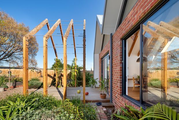 Exterior view of a contemporary brick-clad house opening onto a timber deck and landscaped garden courtyard. A timber pergola with exposed posts and rafters provides a structured outdoor connection, while large dark-framed windows and glazed doors reflect the garden and reveal a bright interior dining area. The scene has clear blue skies, established planting, timber fencing, and warm natural materials.