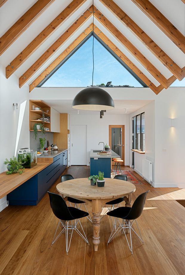 Bright open-plan kitchen and dining area beneath a dramatic pitched vaulted ceiling with exposed timber rafters and beams. The room features white walls, warm timber flooring, dark blue kitchen cabinetry, open timber shelving, a central island, a large black dome pendant, and a round timber dining table with black chairs. A triangular clerestory window brings in abundant daylight, while glazing and a doorway connect to adjacent spaces.