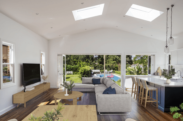 Bright open-plan living room and kitchen with a large grey sectional sofa, timber media console and coffee table, dark timber flooring, and a white kitchen island with bar seating. Expansive glazing and multiple skylights bring in abundant natural light, with a landscaped garden and pool visible outside.