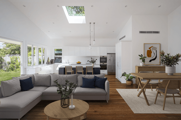 Bright open-plan interior combining a living room, kitchen, and dining area. The space features white cabinetry and walls, warm timber flooring, a large grey sectional sofa with navy cushions, a timber dining table and chairs, skylight, extensive glazing, and contemporary pendant lighting. A framed abstract artwork and potted plants add restrained decorative accents.