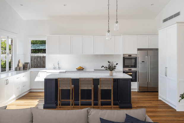 Bright, spacious white kitchen with extensive shaker-style cabinetry, a dark navy island with four woven bar stools, white benchtops and backsplash, integrated double wall ovens, and a stainless-steel refrigerator. Warm timber flooring, pendant lights, and abundant natural light create a refined contemporary setting; the foreground sofa suggests the kitchen opens to a living area.