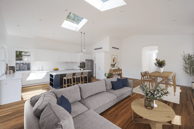 Bright open-plan kitchen, living and dining interior with white cabinetry, a large island, skylights, timber flooring and a grey sectional sofa. The room has abundant natural light, minimalist built-in detailing and a warm neutral palette with dark blue accents.