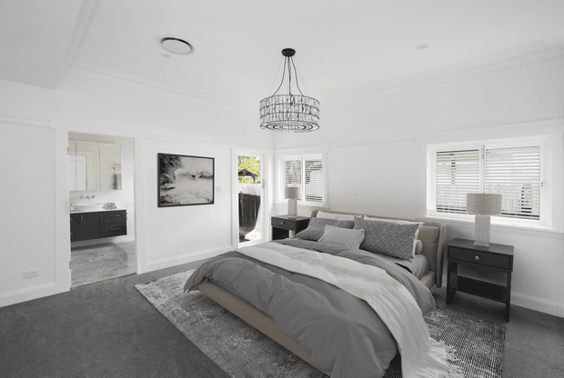 Bright, spacious contemporary bedroom with a large upholstered bed dressed in layered grey and white bedding, matching bedside tables and lamps, a grey rug, white walls with decorative trim, plantation-shuttered windows, and a prominent wire pendant light. An ensuite or bathroom is visible through the open doorway.