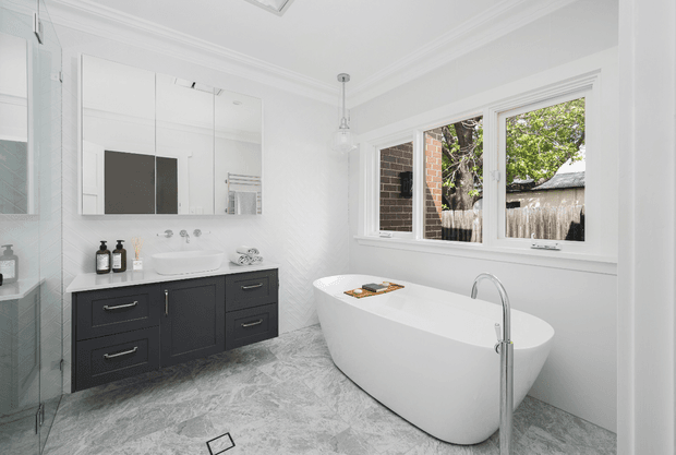Bright contemporary bathroom featuring a freestanding white bathtub beside a large window, a wall-mounted dark vanity with twin basins, mirrored cabinetry, a glass shower enclosure, white textured wall surfaces, and grey marble-look flooring. The room has abundant natural light and a clean, refined finish.