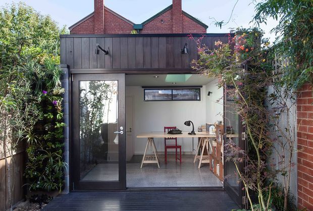 A compact garden studio or workspace opening onto a dark timber deck. The space has a glazed charcoal-framed door, a simple timber workbench with trestle legs, stools, shelving, task lighting and a polished concrete floor. Dense planting surrounds the entrance, while red brick gabled walls are visible behind the dark upper cladding, creating a contemporary backyard extension with strong indoor-outdoor connection.